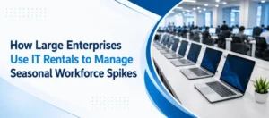 Alt Text: A corporate banner titled "How Large Enterprises Use IT Rentals to Manage Seasonal Workforce Spikes" with clean blue and white geometry. The right side features a curved photo cutout showing long rows of modern business laptops set up on desks in a large enterprise office setting, highlighting bulk IT deployment for temporary staff.
